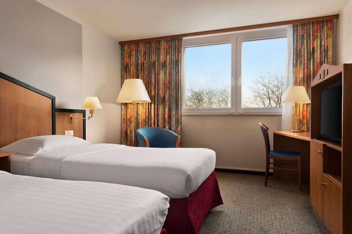 A hotel room with two beds, a desk, a TV, and a window with curtains.