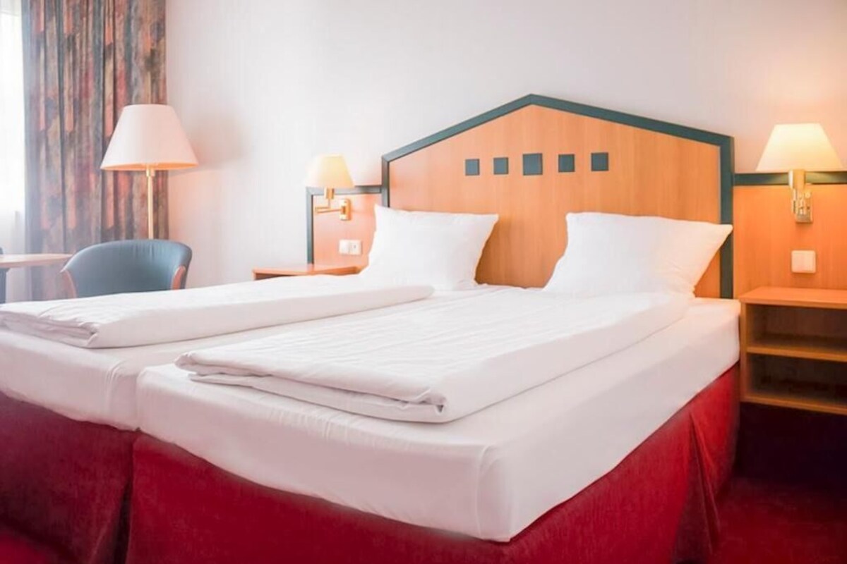 A hotel room with a double bed, two bedside tables with lamps, a chair, and a wooden headboard.