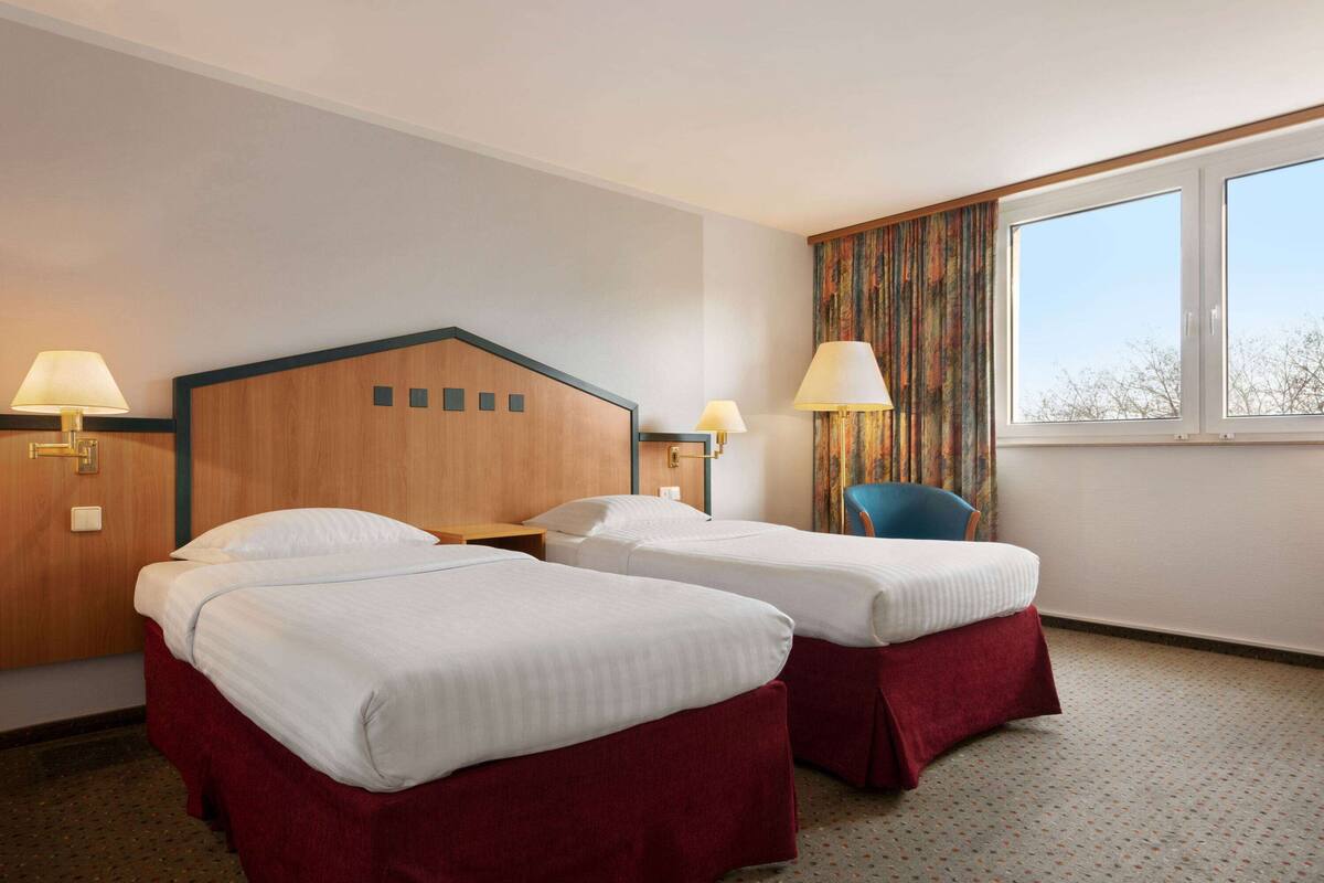 A hotel room with two single beds, a wooden headboard, two bedside lamps, a chair, and a large window.