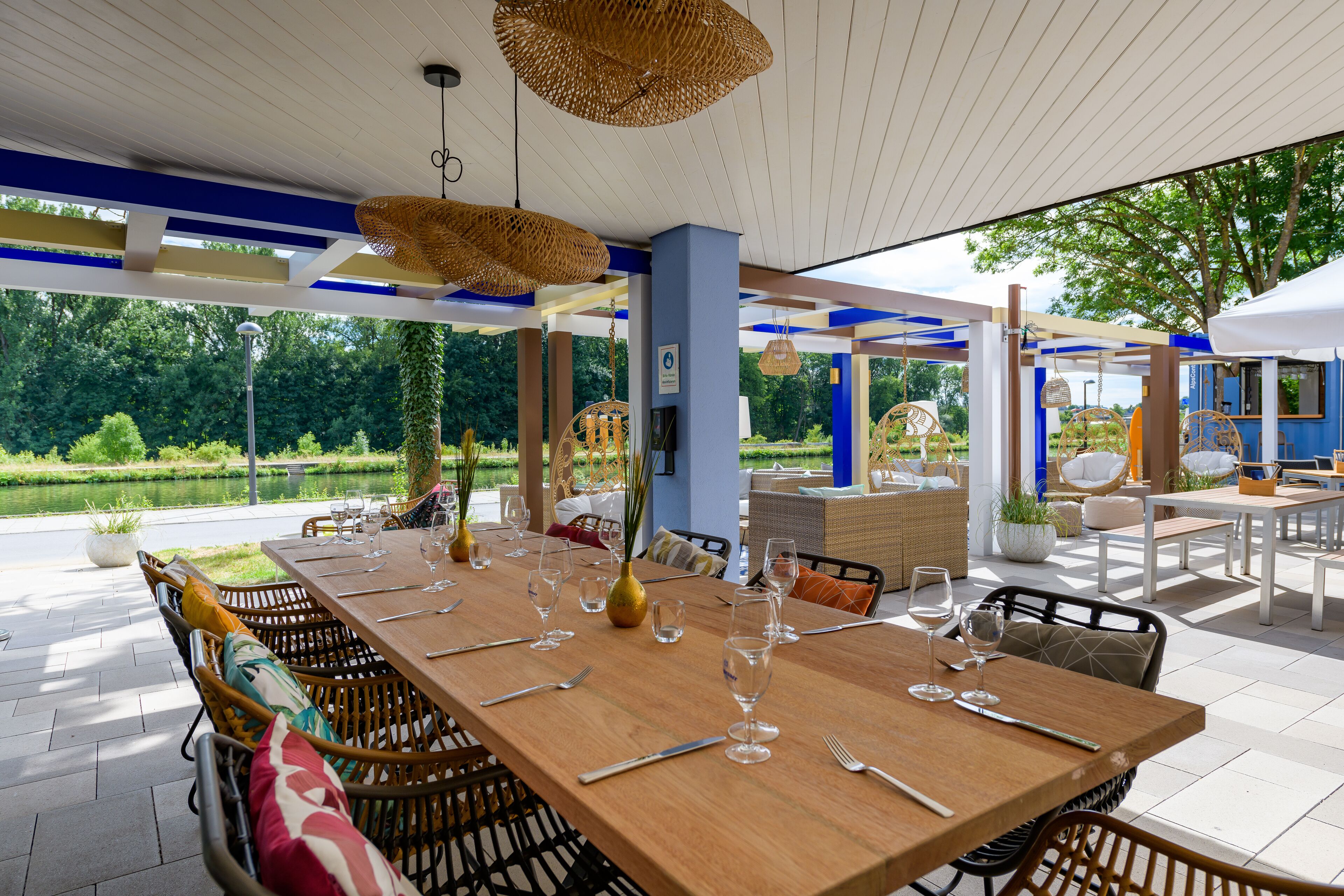 Un área de comedor al aire libre con una larga mesa de madera dispuesta para seis personas, rodeada de sillas de mimbre.