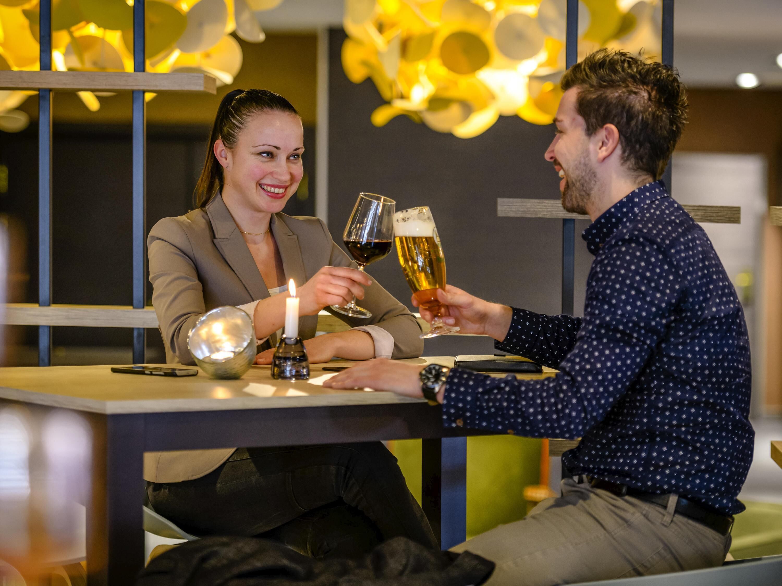 Dos personas brindando con vasos de cerveza en una mesa con una vela, en un ambiente interior moderno con iluminación abstracta.