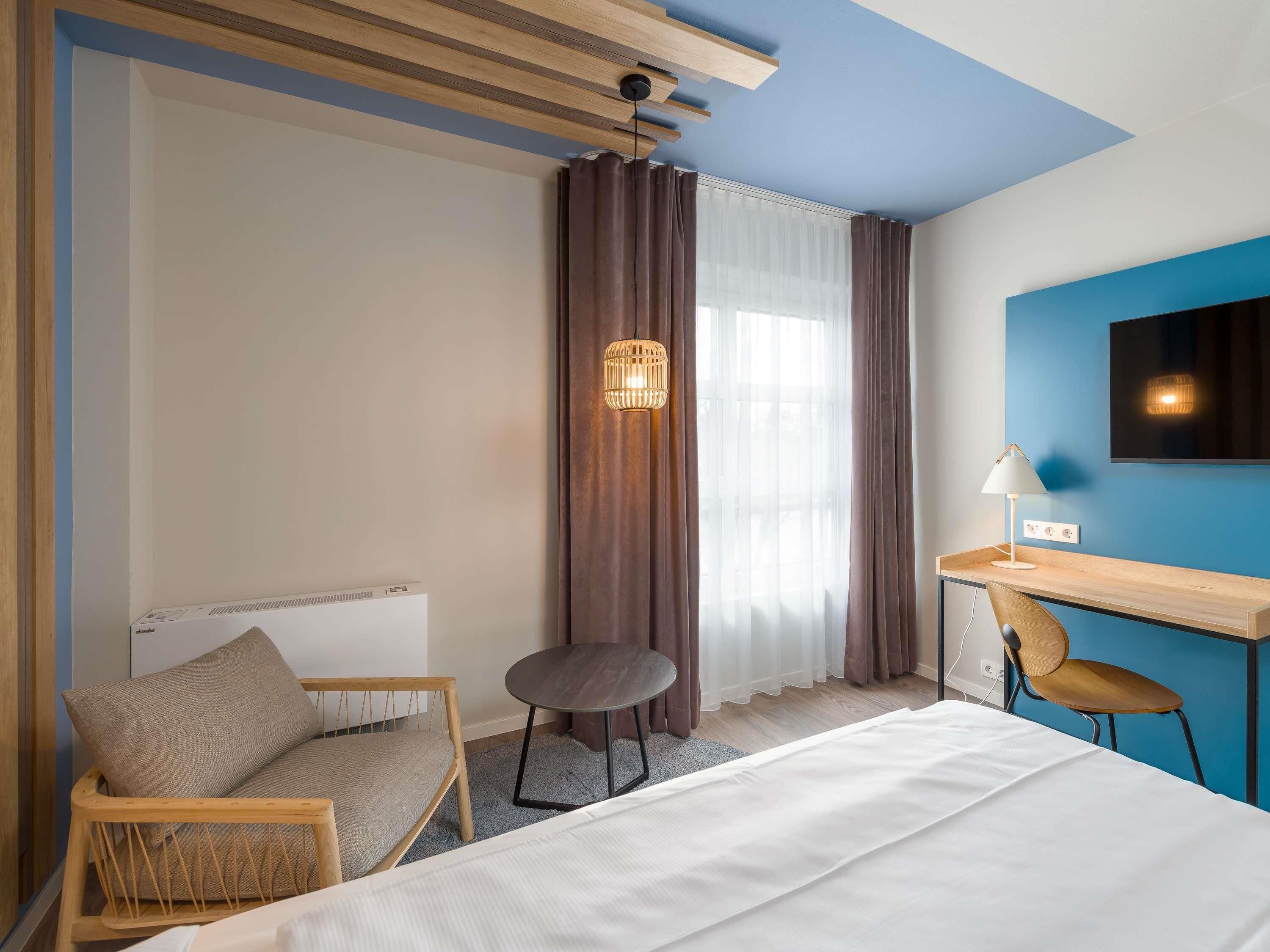 A hotel room with a bed, a desk, a chair, a lamp, a wall-mounted TV, and a window with curtains.