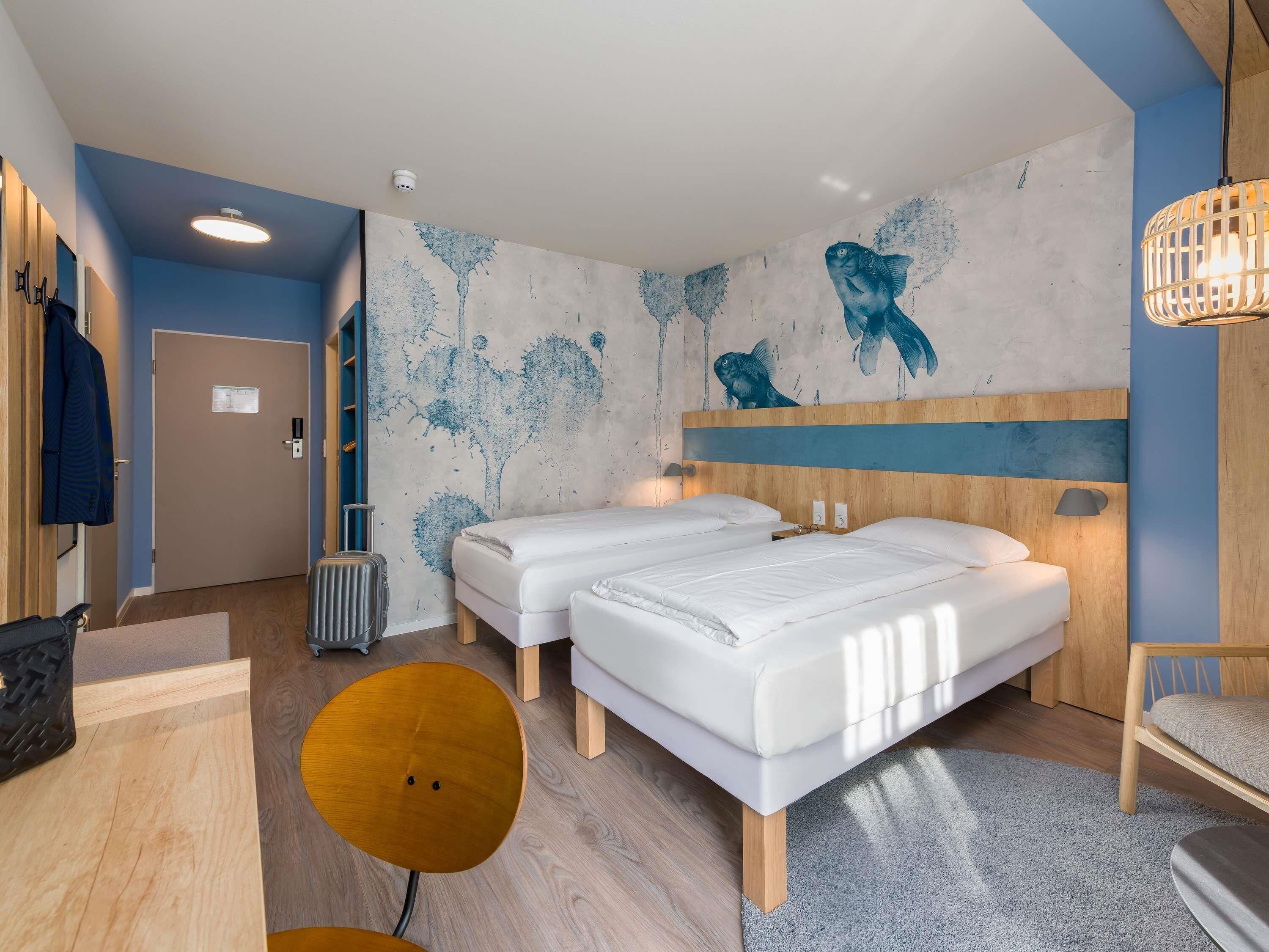 Una habitación de hotel moderna con dos camas, un cabecero de madera, un mural en la pared, una mesa redonda de madera y un radiador.