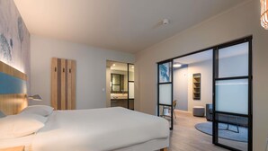 Una habitación de hotel moderna con una cama grande, un escritorio, una silla, una mesita, un espejo y un ventanal corredero que da a un balcón.