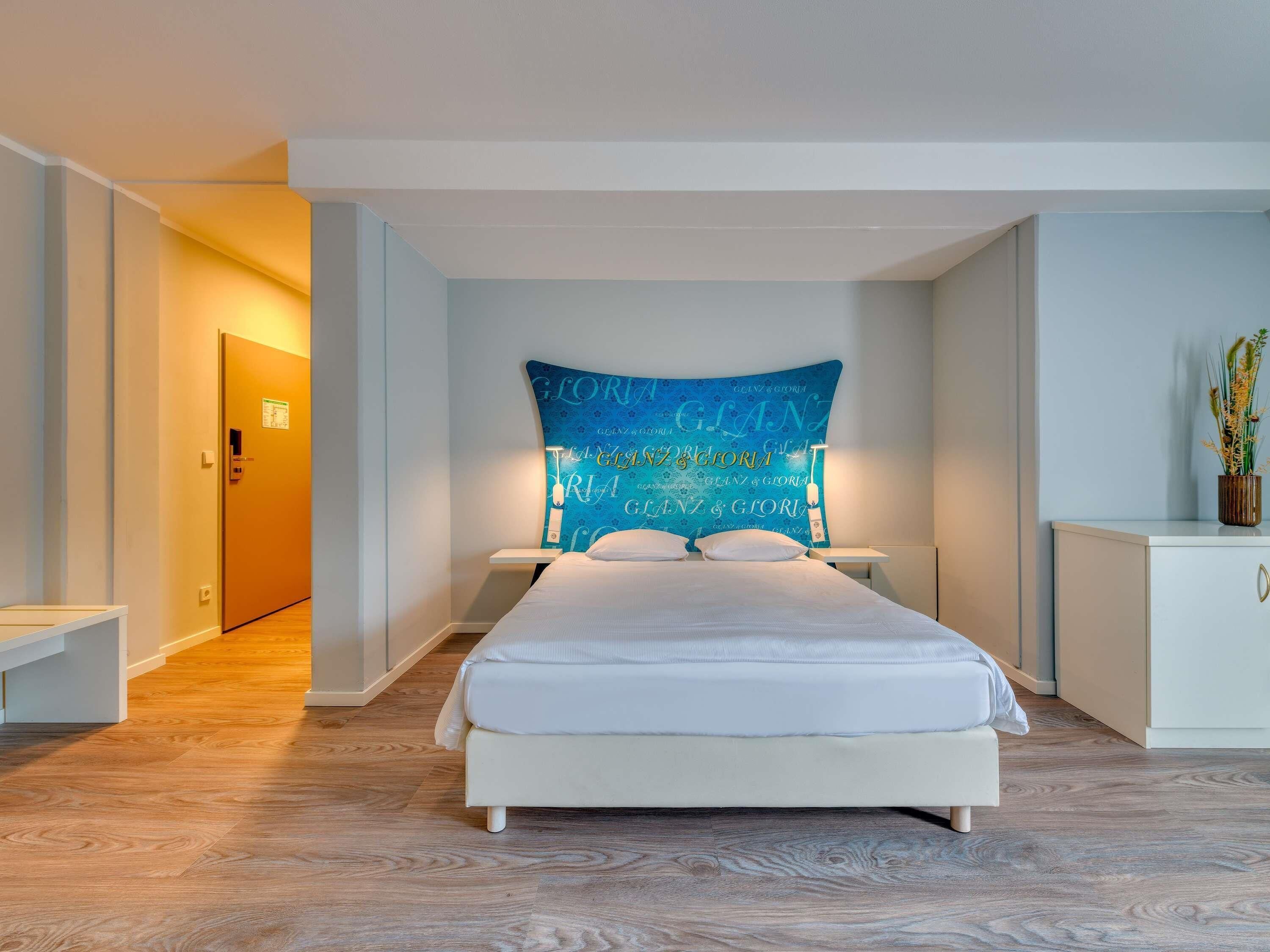 A modern hotel room with a large bed, a nightstand, a bedside table, and a wall-mounted headboard with a blue pattern.