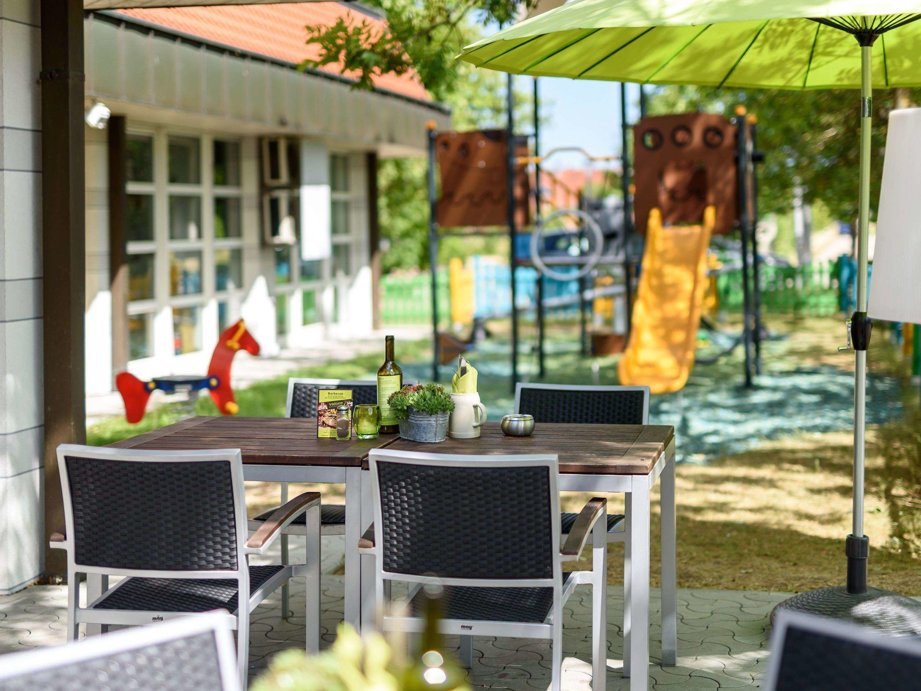 Een buitenterras met een tafel en stoelen, een groene parasol en een speelruimte voor kinderen op de achtergrond.