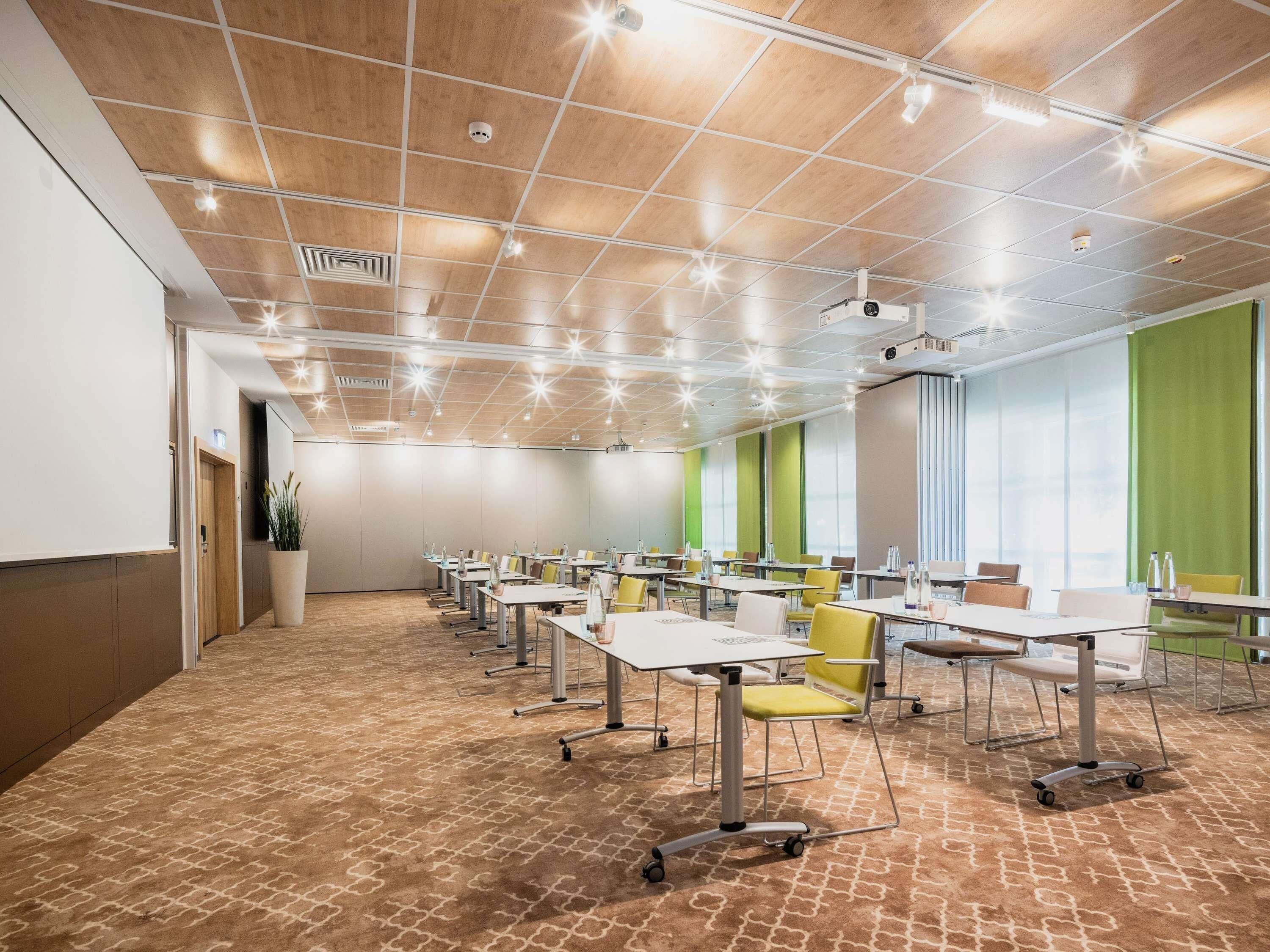 Une salle de conférence dotée d’un plafond à motifs géométriques, de plusieurs tables entourées de chaises et d’une grande fenêtre ornée de rideaux verts.