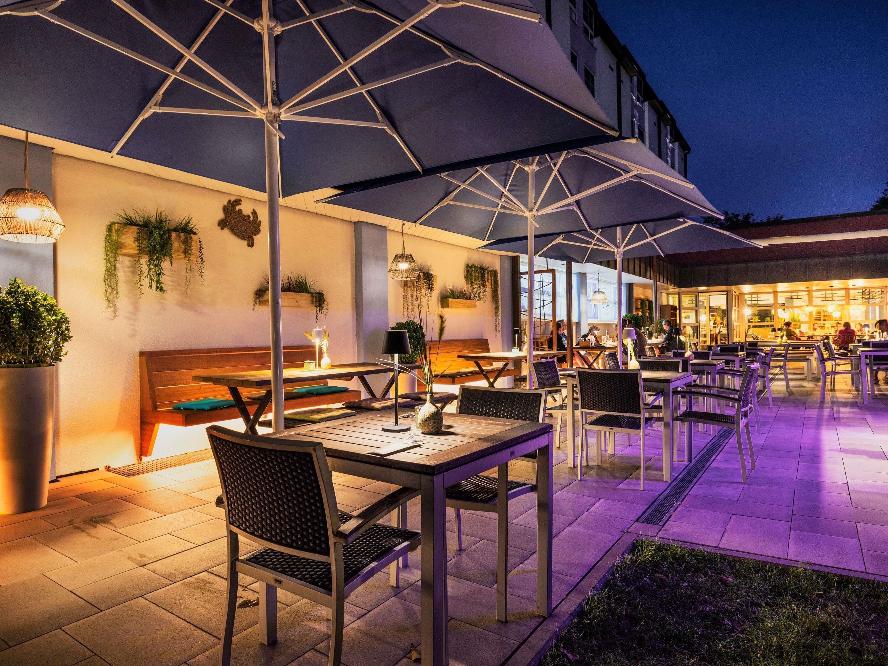 An outdoor dining area with tables and chairs, umbrellas, and a building in the background.