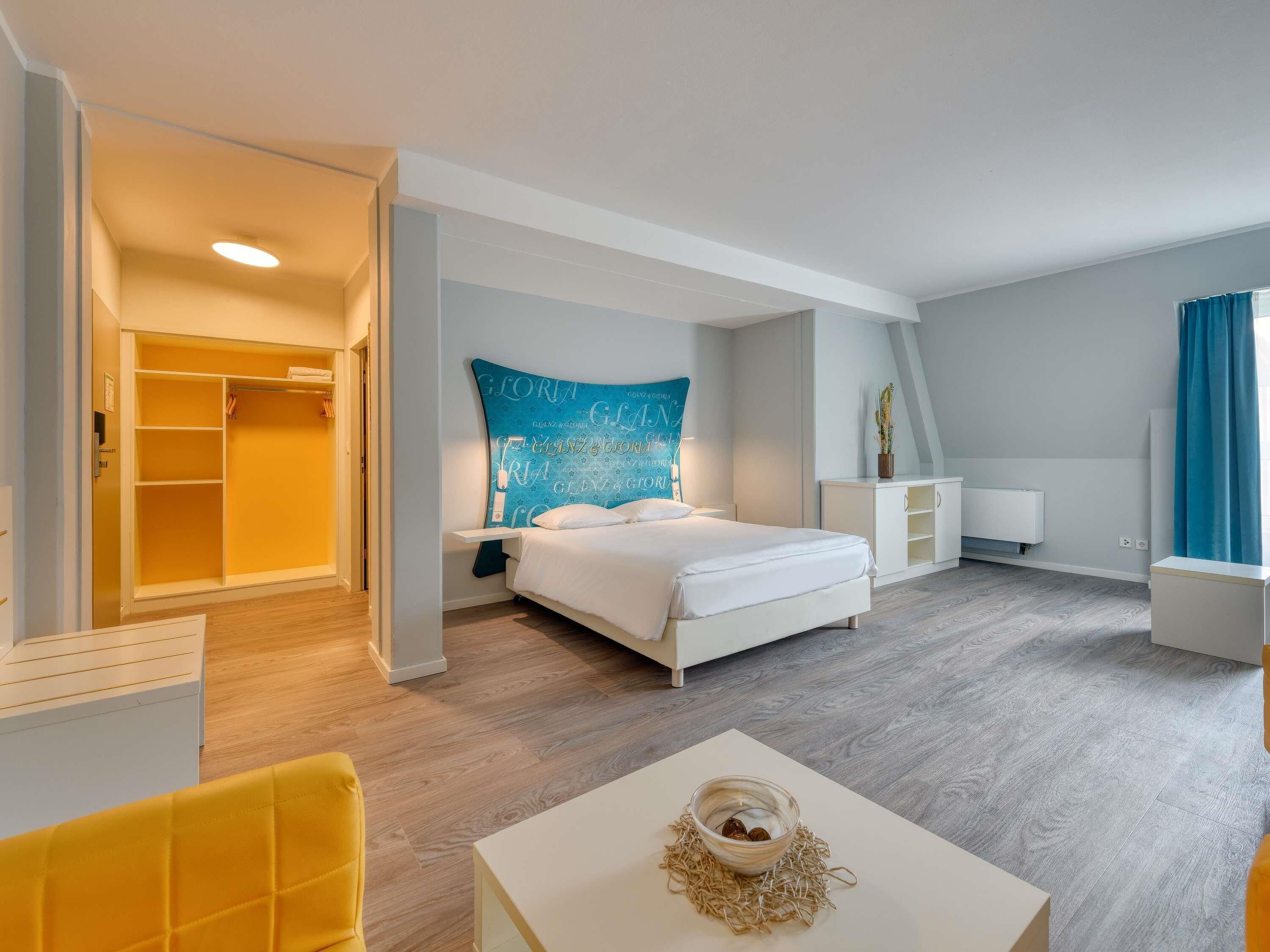 A hotel room with a bed, bedside table, a nightstand, a wardrobe, a seating area with a chair, a small table, and a decorative bowl.