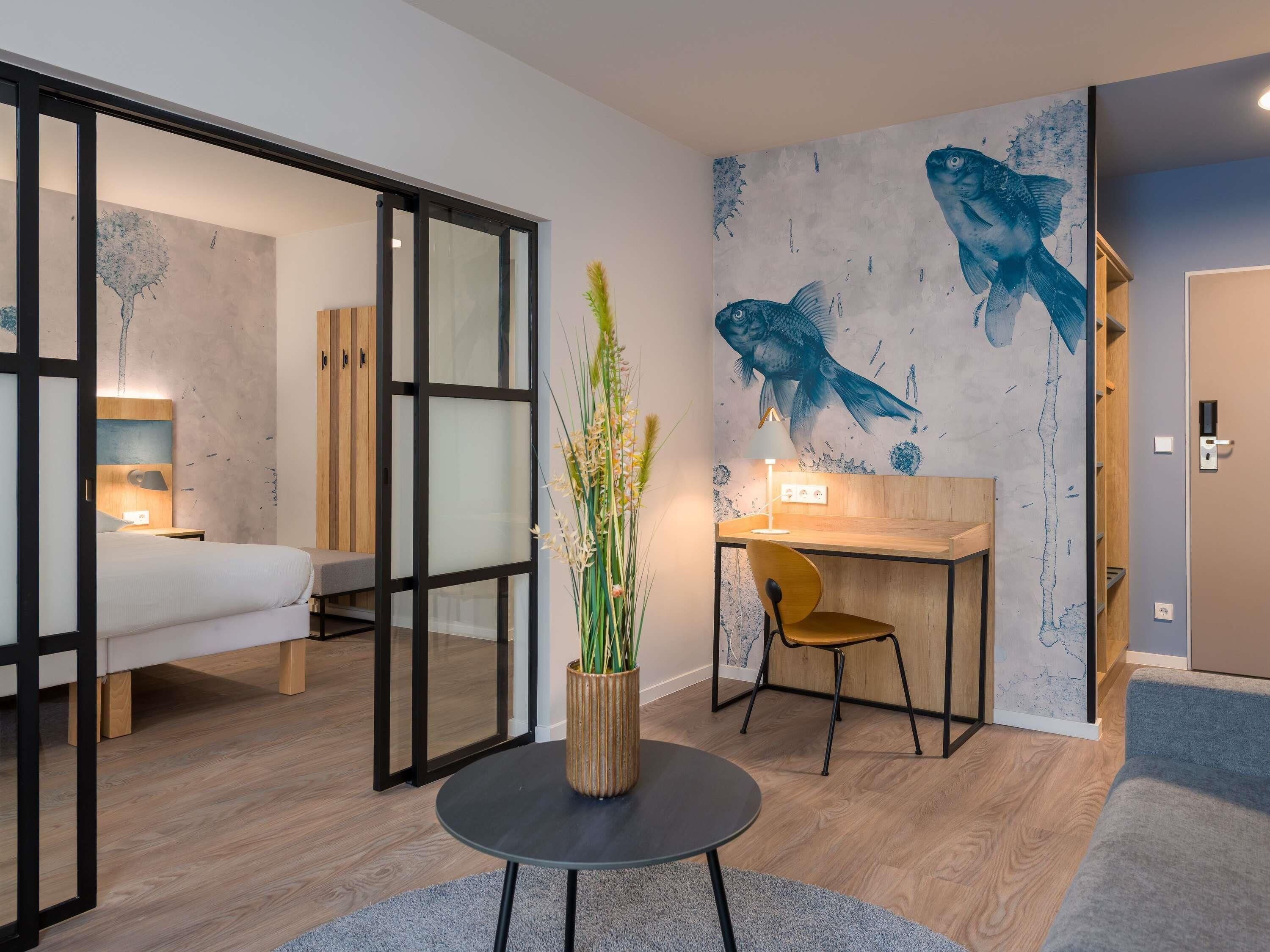 A modern hotel room with a bed, a desk, a chair, a round table with a plant, and a wall with fish artwork.