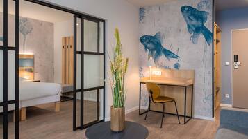 A modern hotel room with a bed, a desk, a chair, a round table with a plant, and a wall with fish artwork.