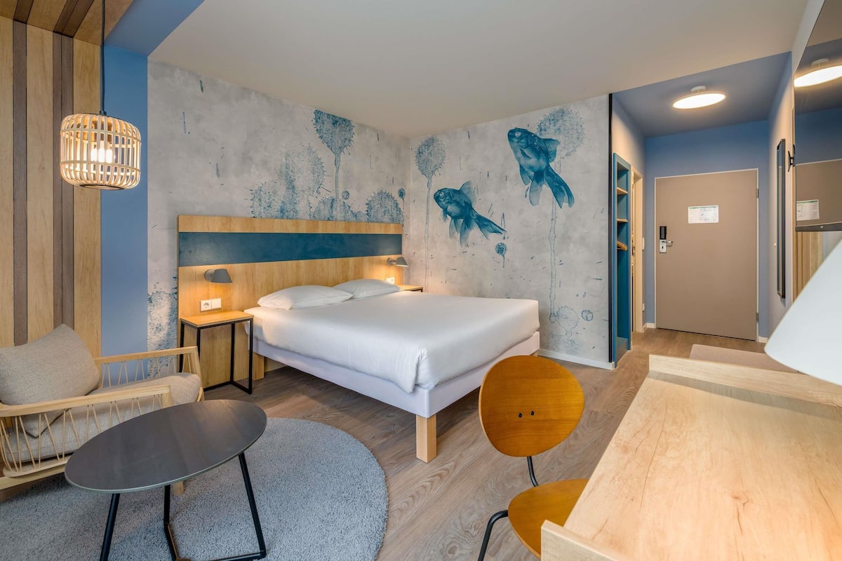 A modern hotel room with a bed, a wooden headboard, a wall mural, a round table, two chairs, a small table, and a door.