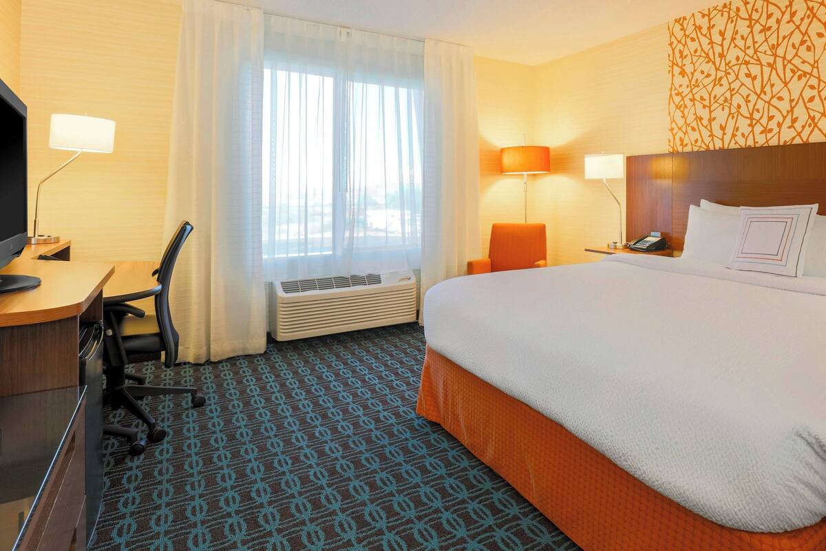 A hotel room with a bed, desk, chair, television, lamps, and a patterned carpet.