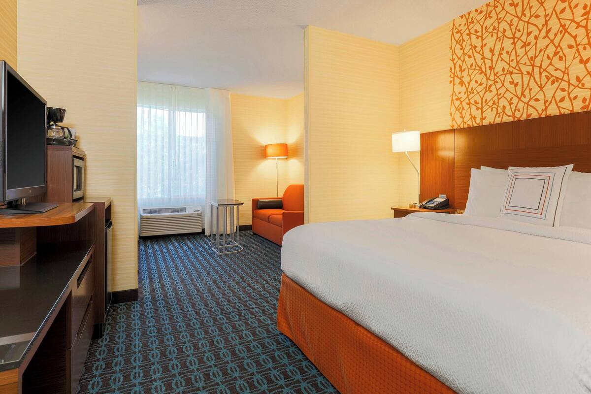 A hotel room with a bed, a desk with a television, a chair, a lamp, and a window with blinds.