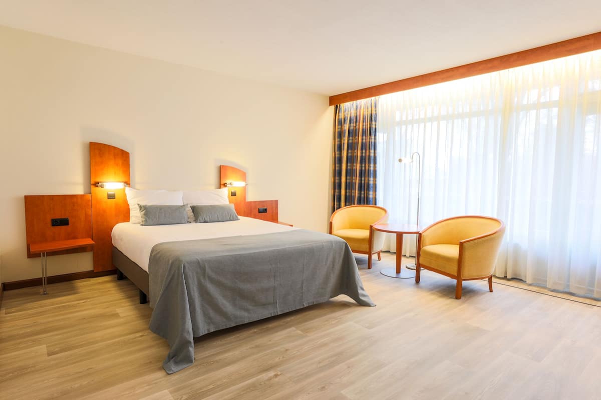 A hotel room with a bed, two nightstands, a bedside table, a chair, and a window with curtains.