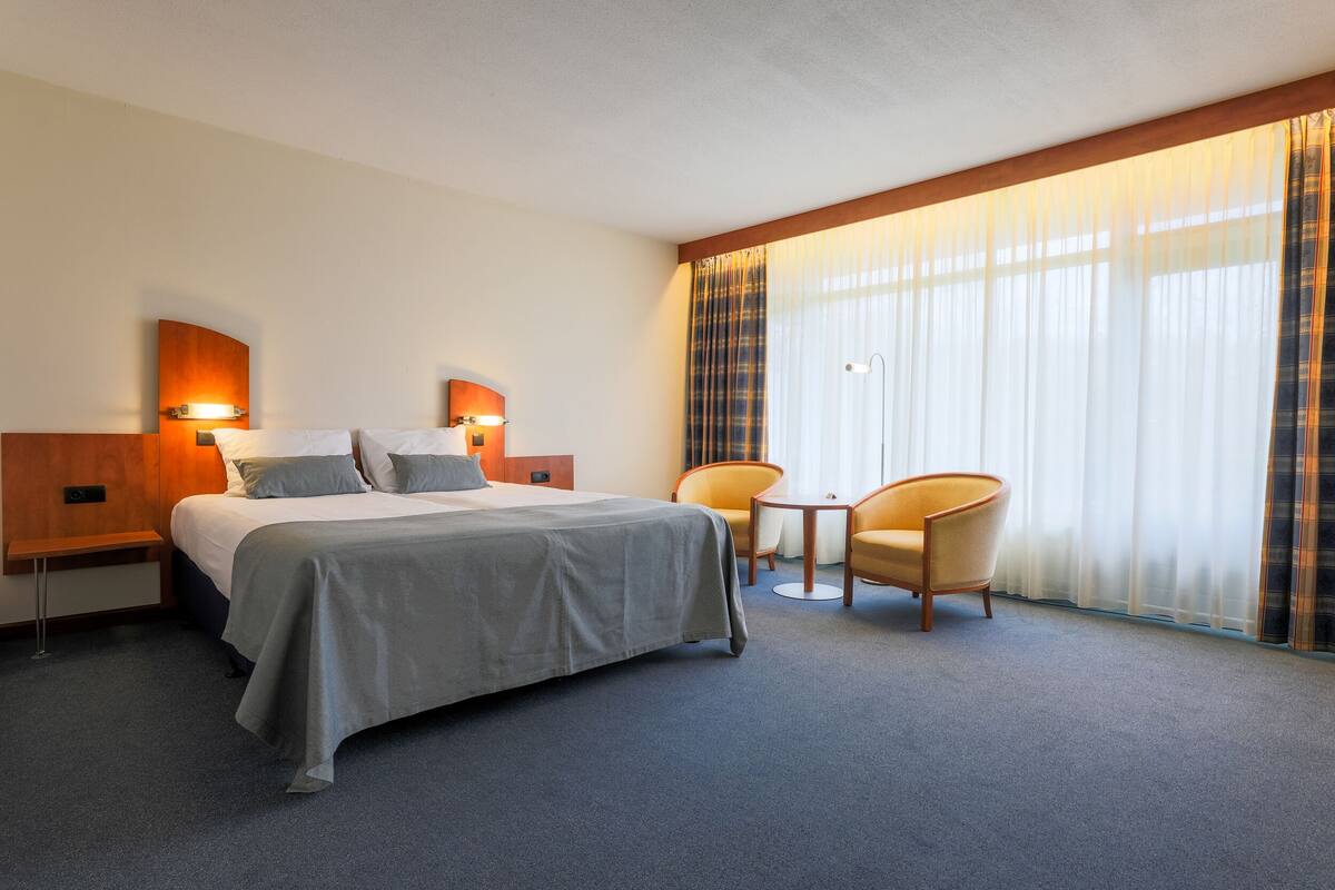 A hotel room with a bed, two bedside tables, a chair, a small table, and a large window with curtains.
