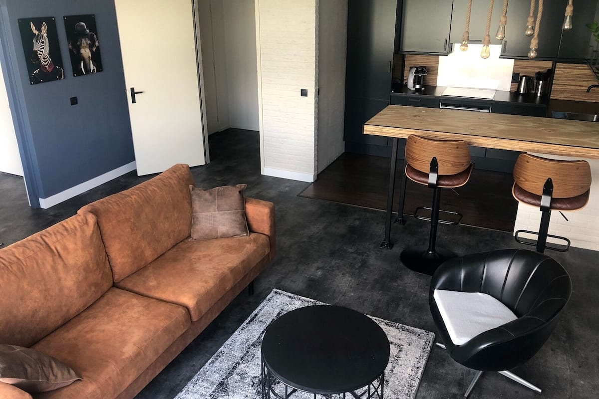 A modern living room with a brown sofa, a black round table, and a black chair. There is a kitchen area with a wooden countertop and two bar stools.