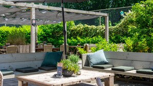 An outdoor seating area with wooden benches and a table, surrounded by greenery and a pergola with a canopy. - Fletcher Hotel - Restaurant De Reiskoffer (Bosschenhoofd)
