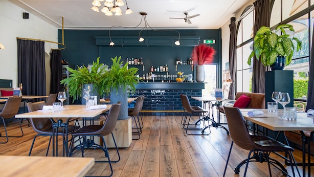 A restaurant with wooden flooring, tables with chairs, a bar area with shelves of bottles, and potted plants. - Fletcher Hotel - Restaurant De Reiskoffer (Bosschenhoofd)