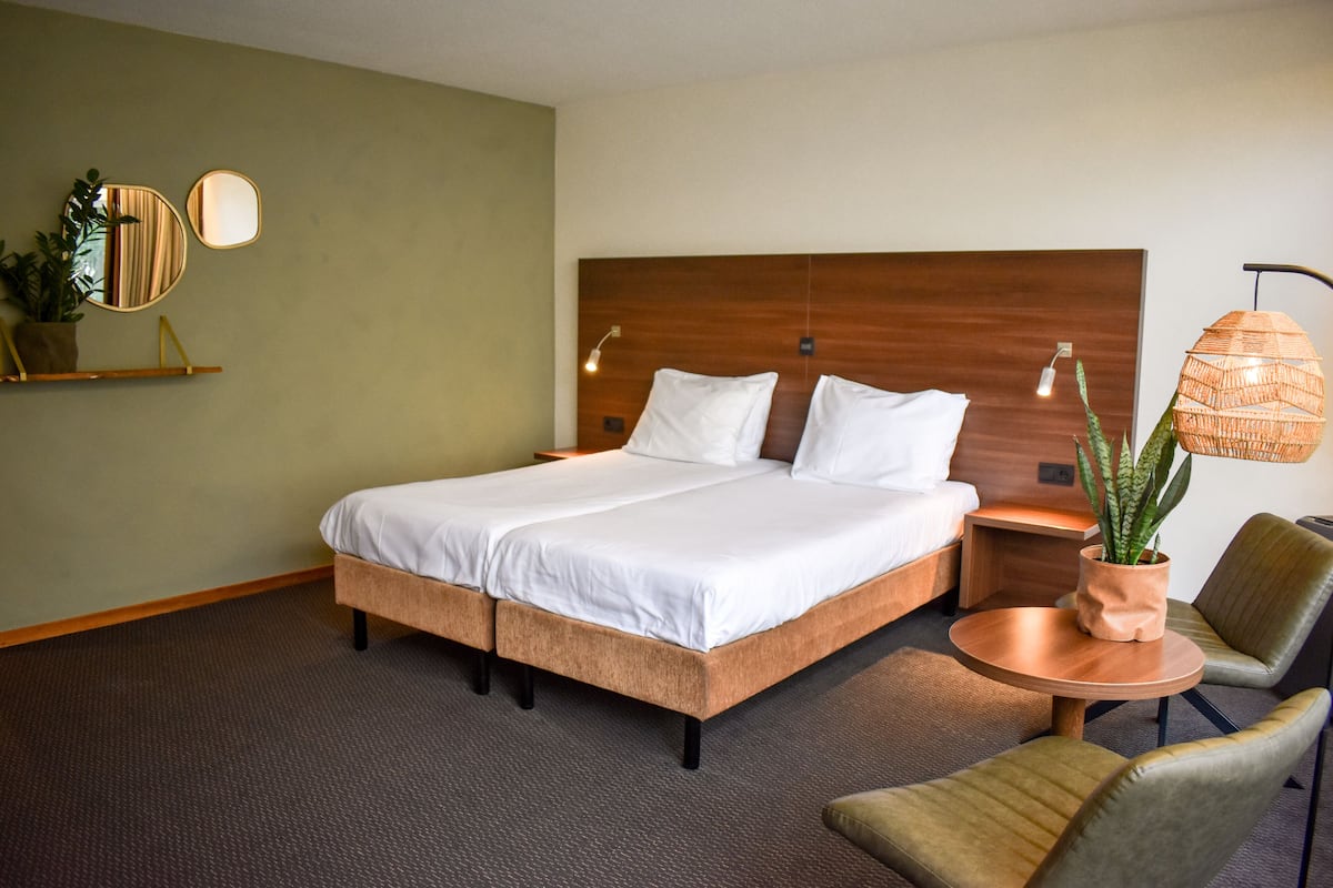 A hotel room with a large bed, a bedside table, a round coffee table, two chairs, a potted plant, and a wall-mounted mirror.