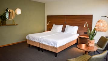 A hotel room with a large bed, a bedside table, a round coffee table, two chairs, a potted plant, and a wall-mounted mirror.