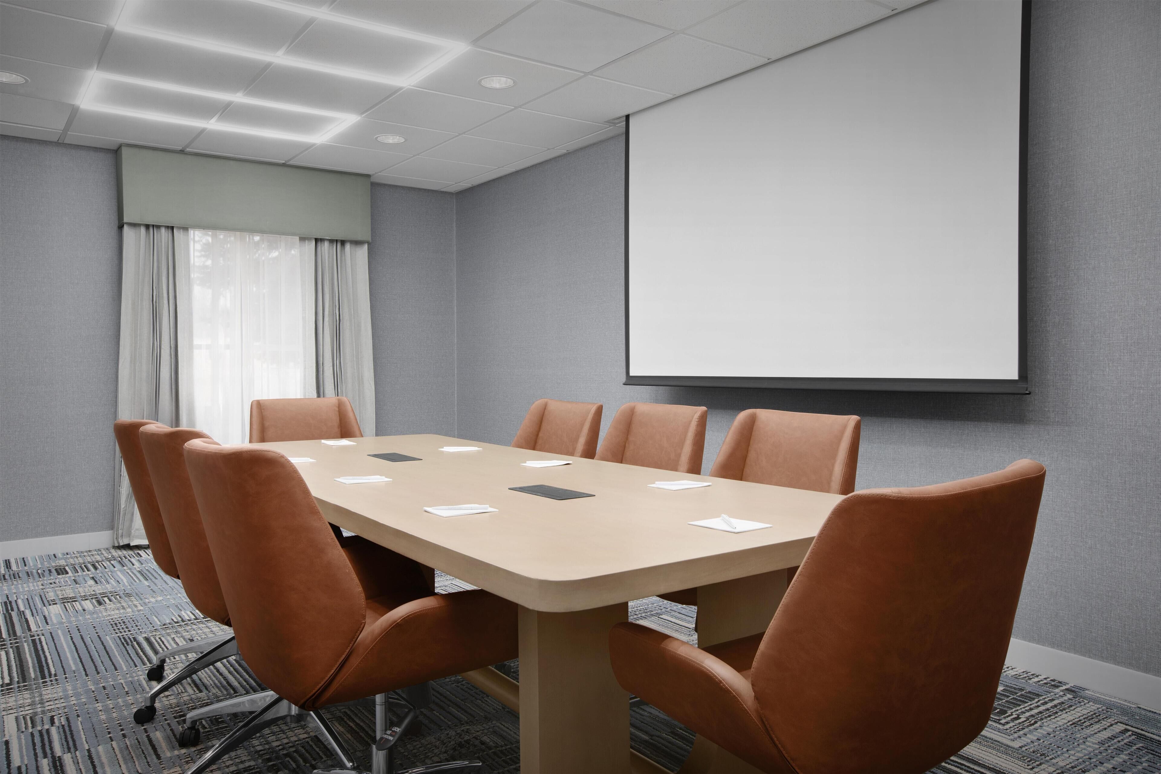 Une salle de conférence équipée d’une grande table, de six chaises, d’un écran de projection et d’un sol recouvert de moquette.
