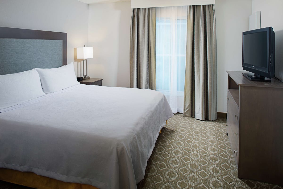 Una habitación de hotel con una cama grande, un televisor en una cómoda, una mesita de noche con lámpara y una ventana con cortinas.
