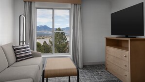 Ett hotellrum med en soffa, en tv på ett träställ, ett fönster med utsikt över bergen och ett litet bord.