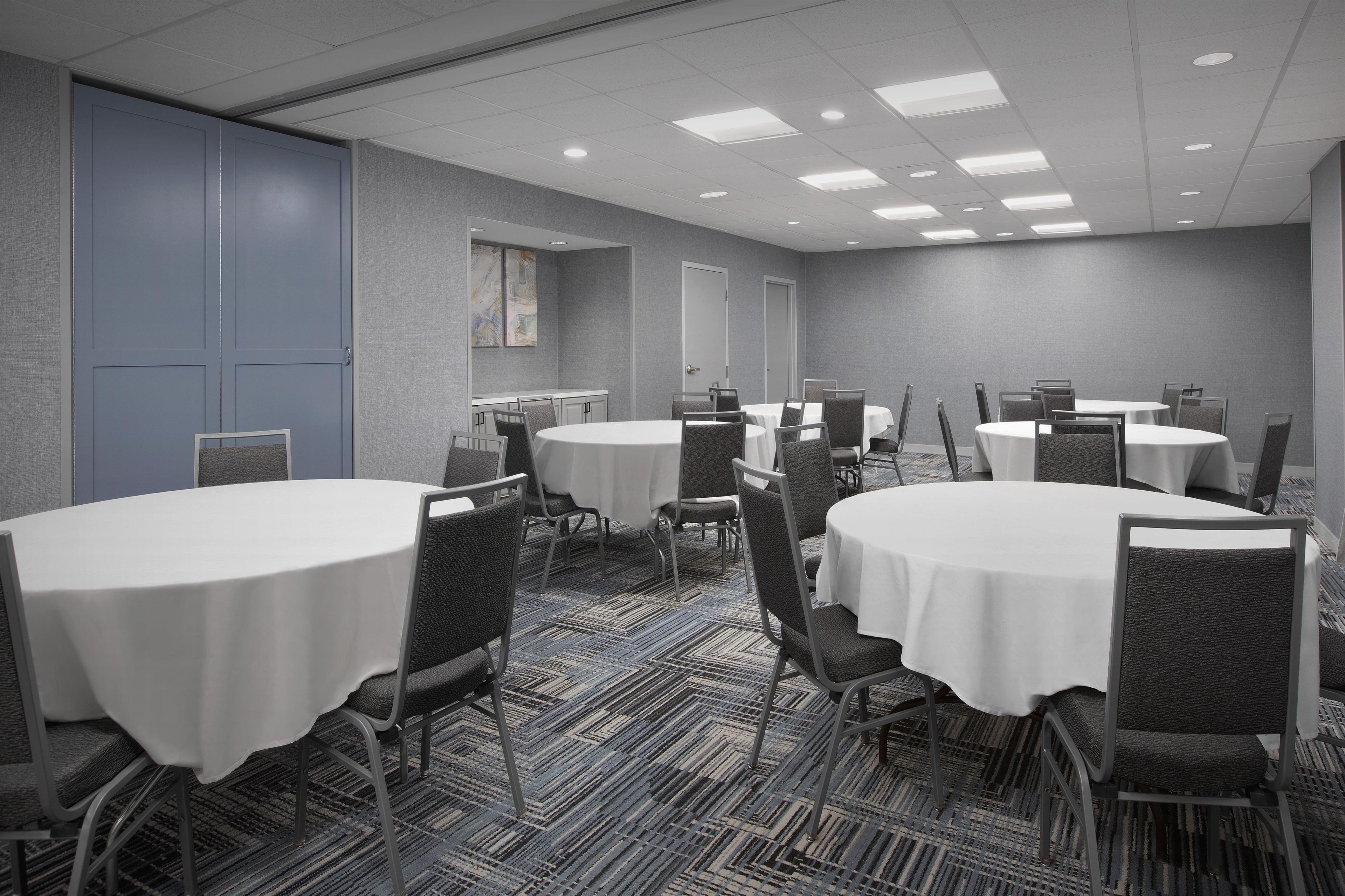 A conference room with multiple tables and chairs, carpeted flooring, and a plain wall with a door and a window.