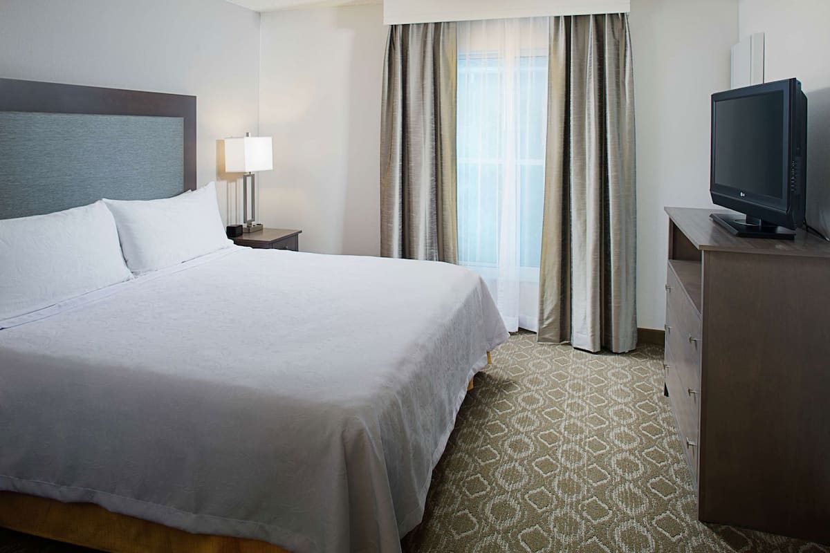 Una habitación de hotel con una cama grande, un televisor en una cómoda, una mesita de noche con lámpara y una ventana con cortinas.