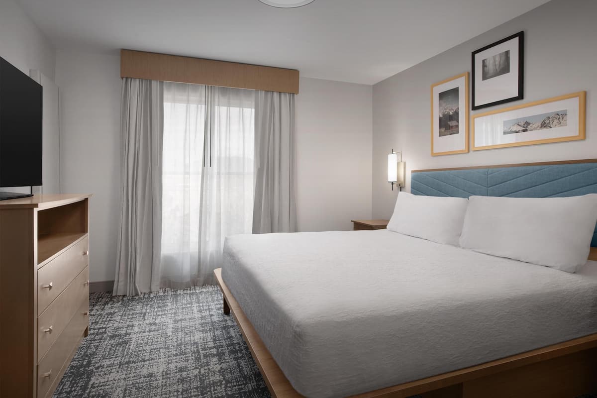 Habitación de hotel con cama, televisión, escritorio, silla, lámpara y ventana con cortinas.