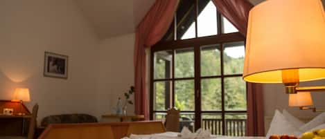 A hotel room with a large window, a bed with white linens, a chair, a table with a lamp, and a fruit bowl on the side.
