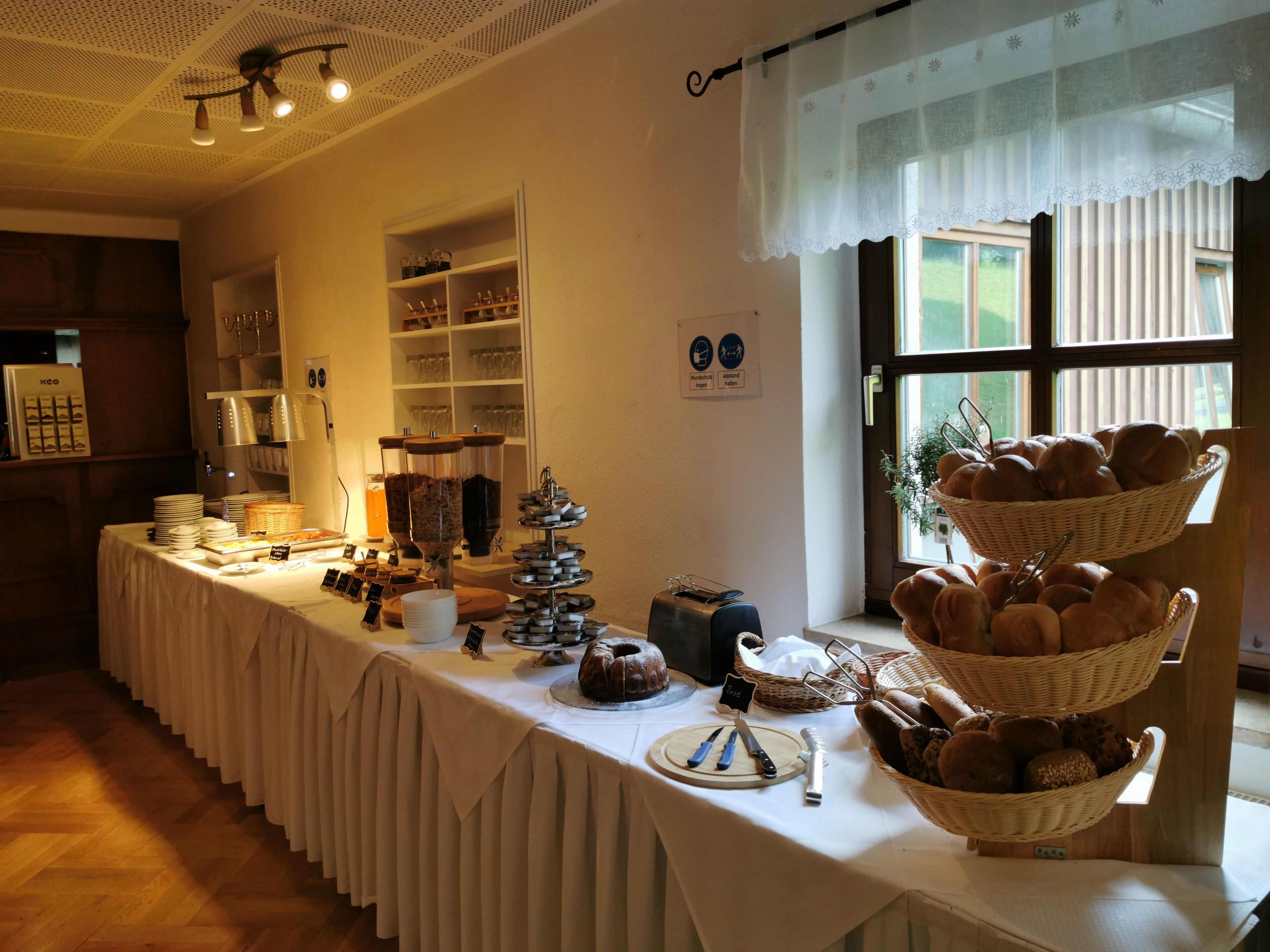 Ein reichhaltiges Buffet mit verschiedenen Speisen und Besteck auf einer Tischplatte, umgeben von Regalen mit Glaswaren und einem Fenster mit Blick.