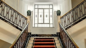 Una imponente escalera de mármol con alfombra roja, que conduce a un amplio ventanal con vidrieras decorativas.