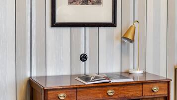 A wooden desk with drawers, a framed picture above it, a small round ottoman, and a gold desk lamp.