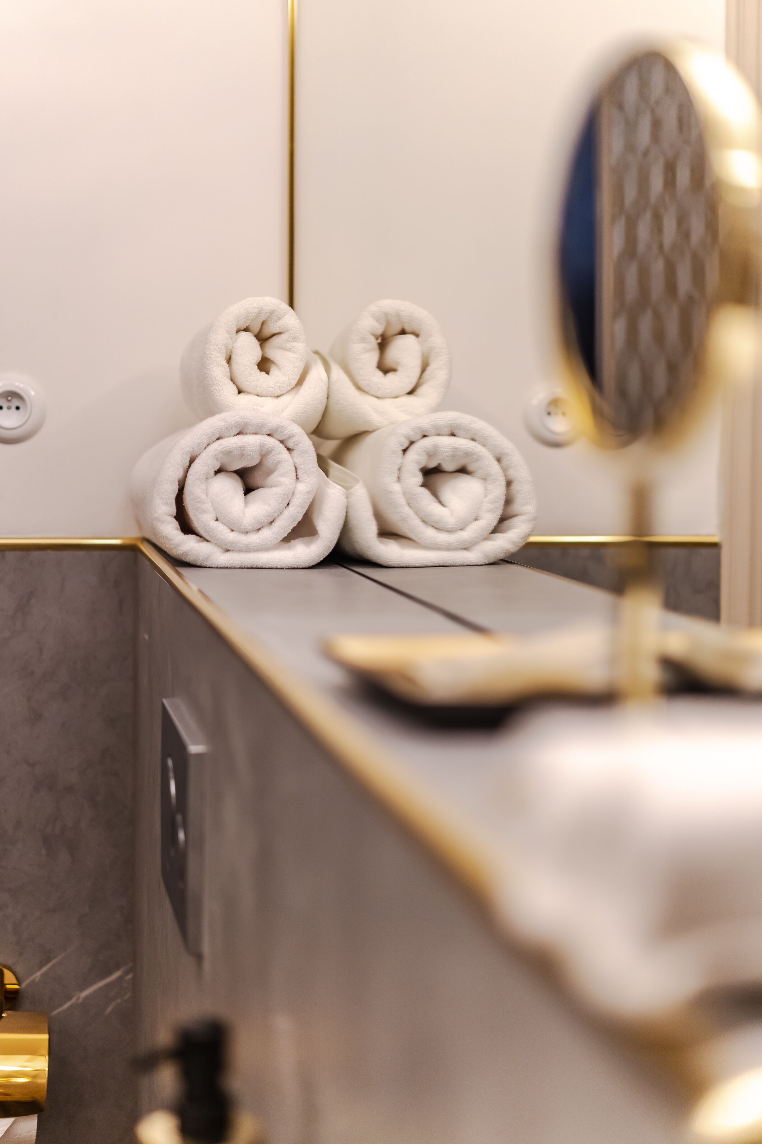 Un baño con un perchero que sostiene tres toallas enrolladas, un espejo y un lavabo dorado.