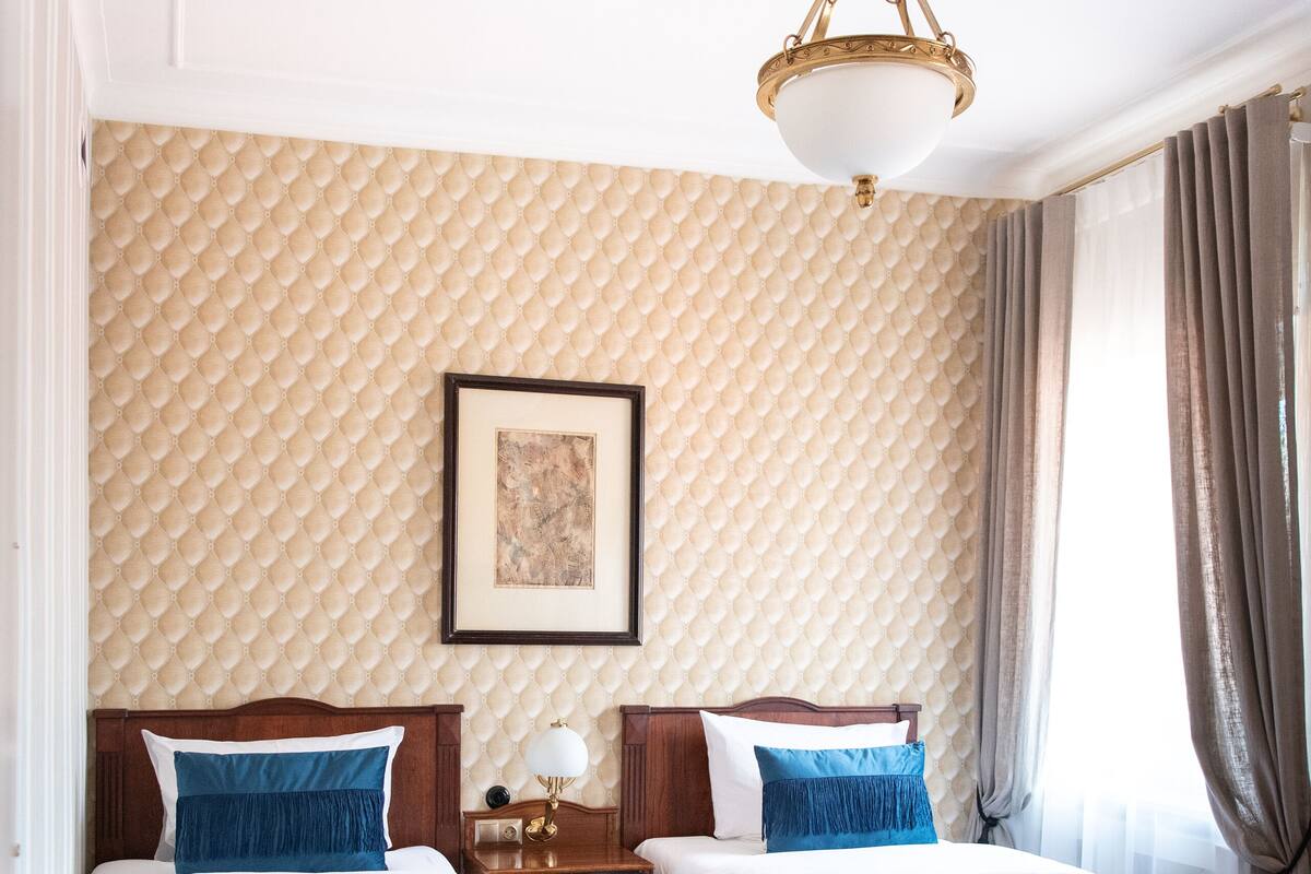 A hotel room with two beds, a chandelier, a framed picture on the wall, and a window with curtains.