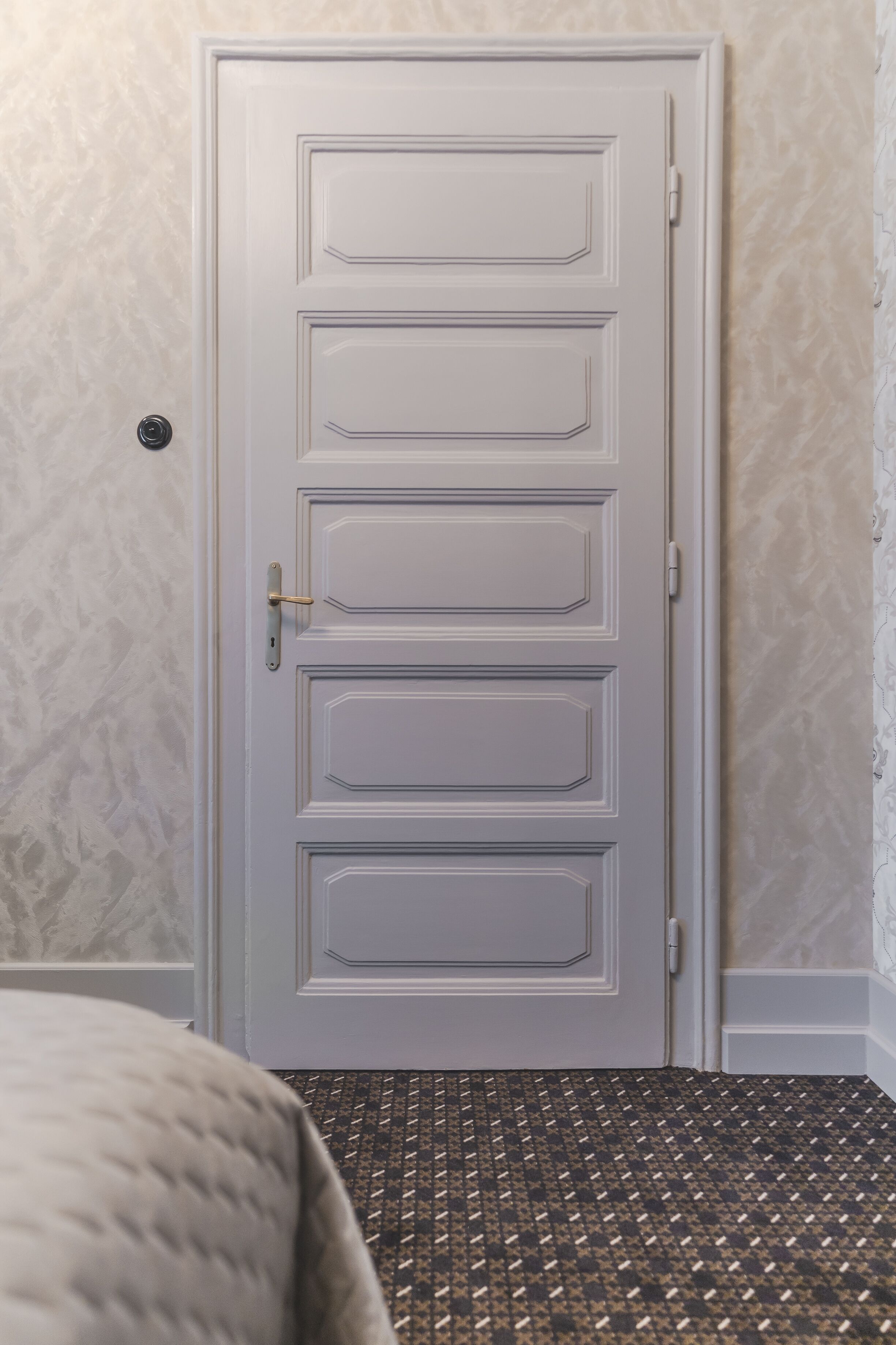 A closed door with a simple handle and a keyhole, set in a room with patterned carpet and textured wallpaper.