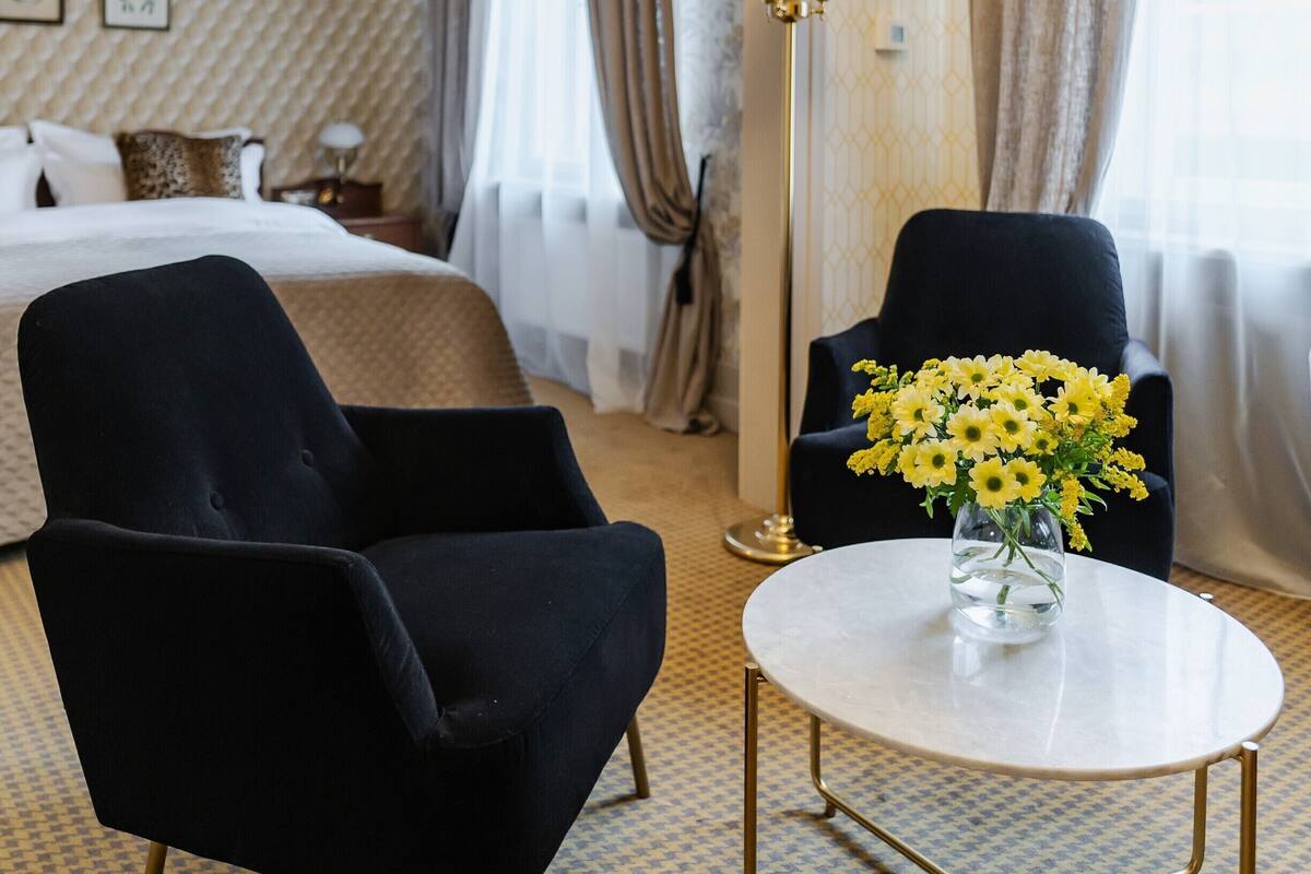A hotel room with a bed, two armchairs, a small round table with a vase of flowers, a floor lamp, and patterned carpet.