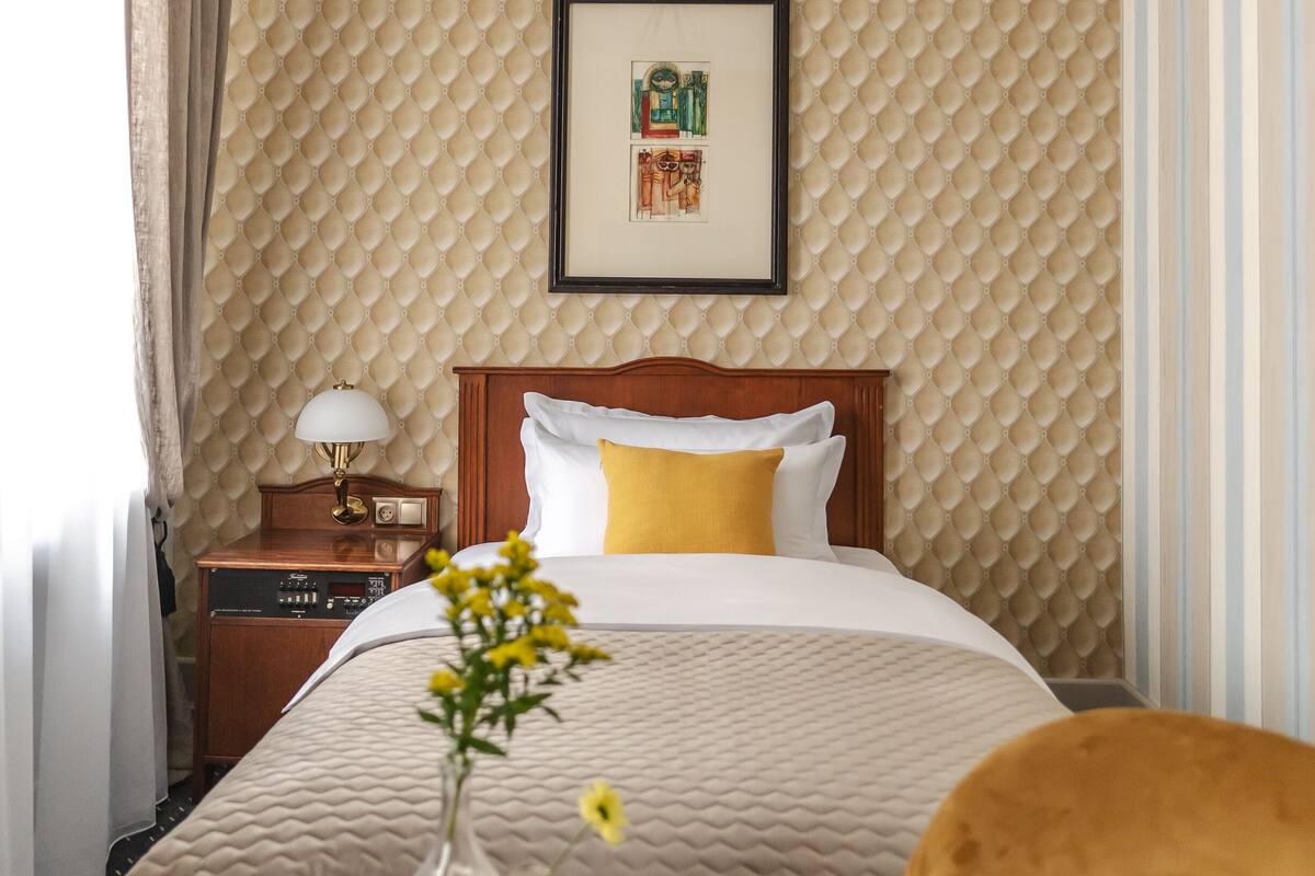A hotel room with a bed, a bedside table with a lamp, a framed picture on the wall, and a window with curtains.