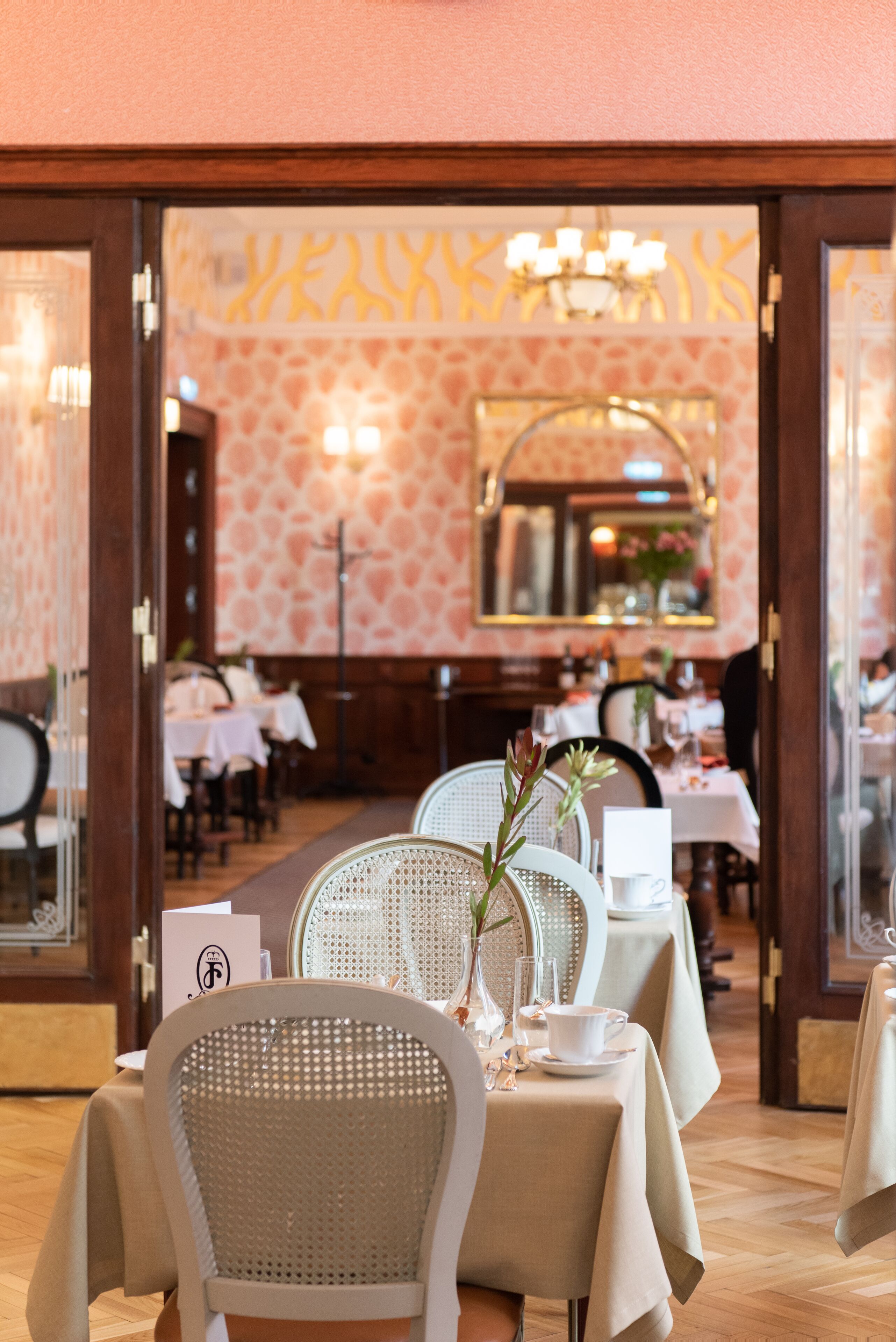 L’intérieur d’un restaurant, avec des tables dressées pour le service, un miroir au mur et un papier peint à motifs.