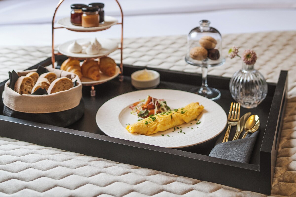 A breakfast tray with pastries, a plate of food, and a glass jar on a bed with a patterned headboard.