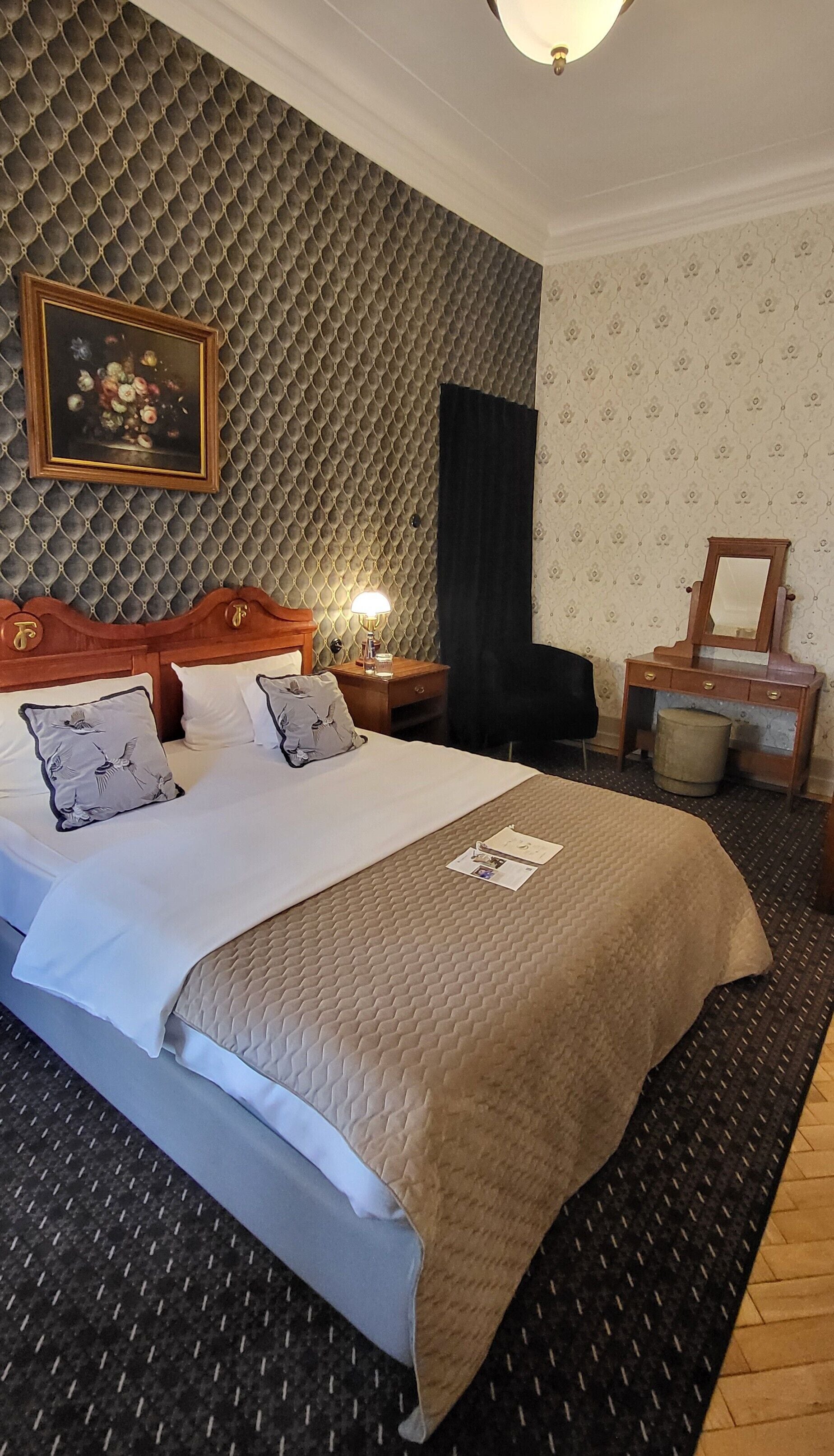 A hotel room with a large bed, a wooden headboard, a nightstand with a lamp, a dresser with a mirror, and a painting on the wall.