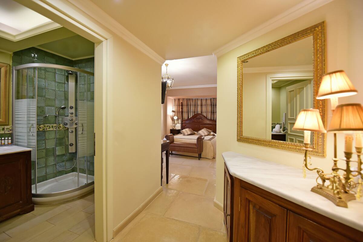 A bathroom with a glass-enclosed shower, a vanity with a mirror, and a bed in the background.