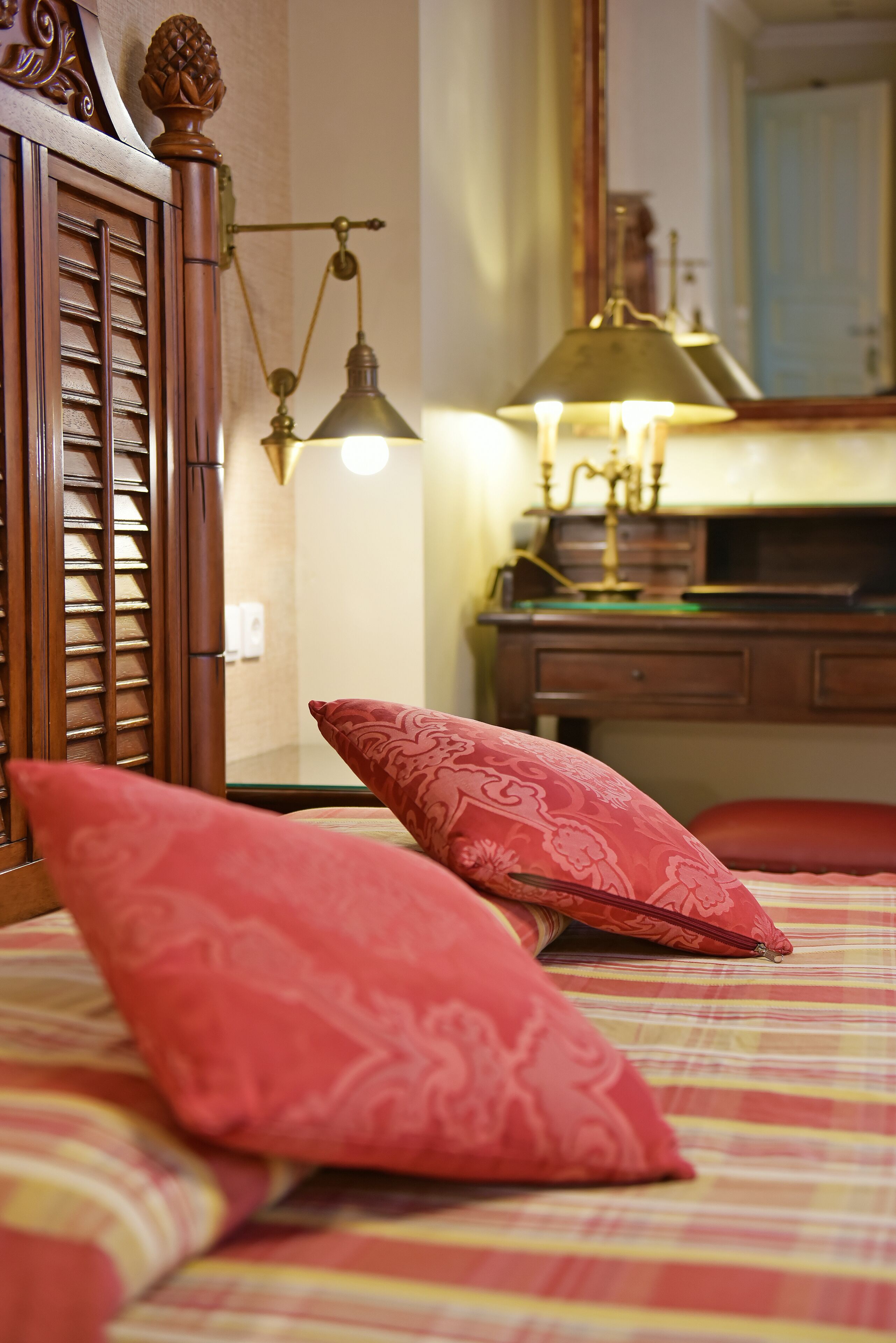 Une chambre à coucher avec un lit, des coussins à motifs rouges, une tête de lit en bois, un bureau avec une lampe et un miroir.