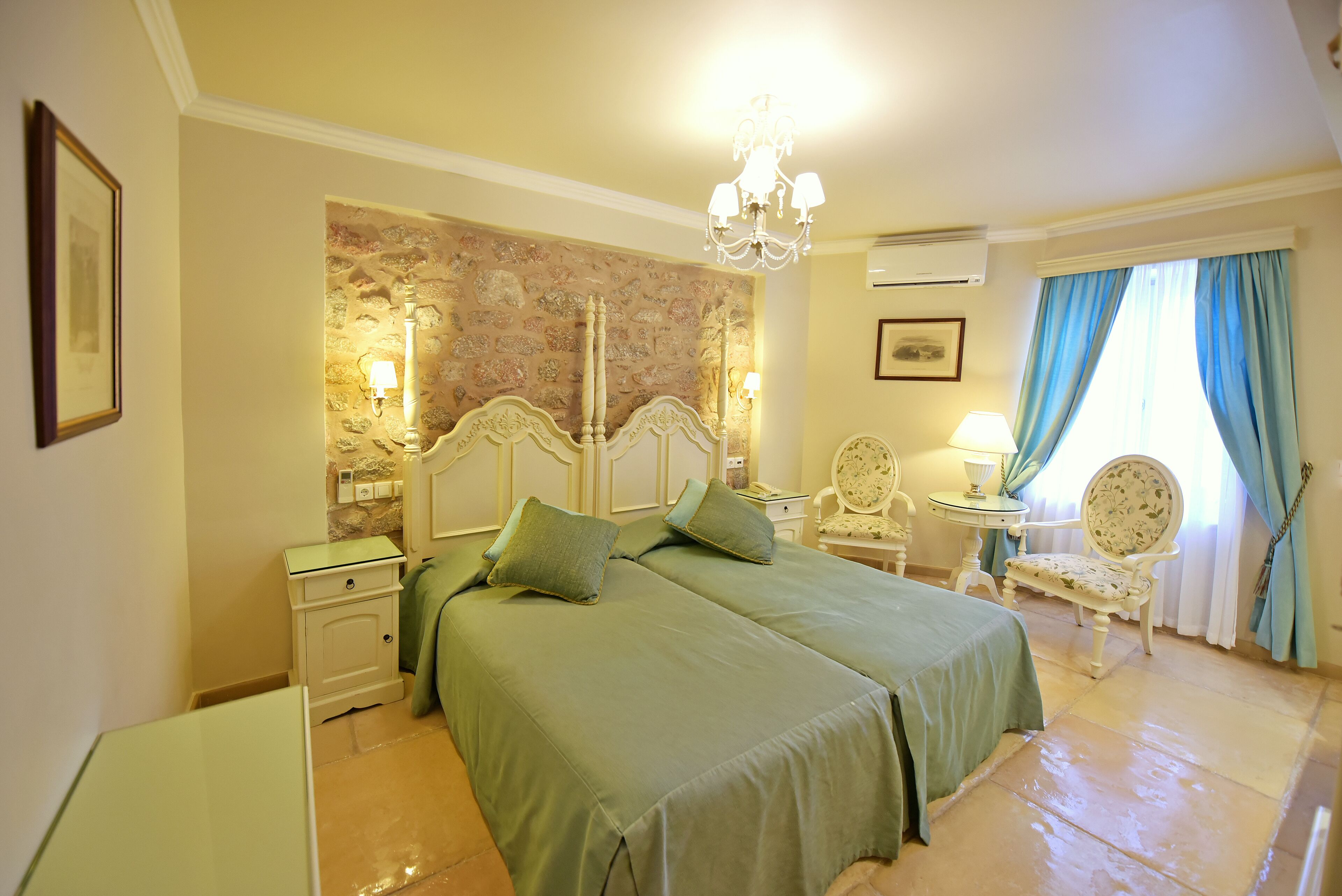 A hotel room with two beds, a stone accent wall, a chandelier, a small table with a lamp, a chair, and a window with curtains.