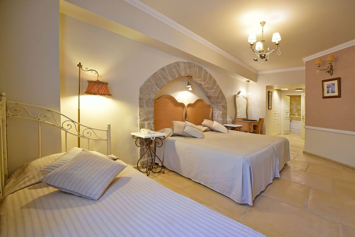 A hotel room with two beds, a stone archway, a chandelier, and a small table with a lamp.