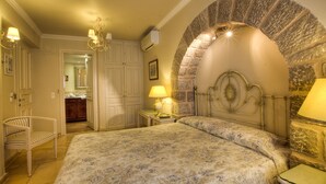 A bedroom with a large bed, a stone archway, a chandelier, and a small table with a lamp. - Angelica Boutique Hotel (Hydra)