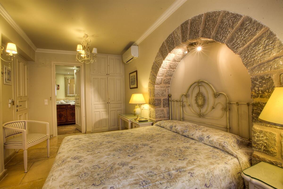 A bedroom with a large bed, a stone archway, a chandelier, and a small table with a lamp.