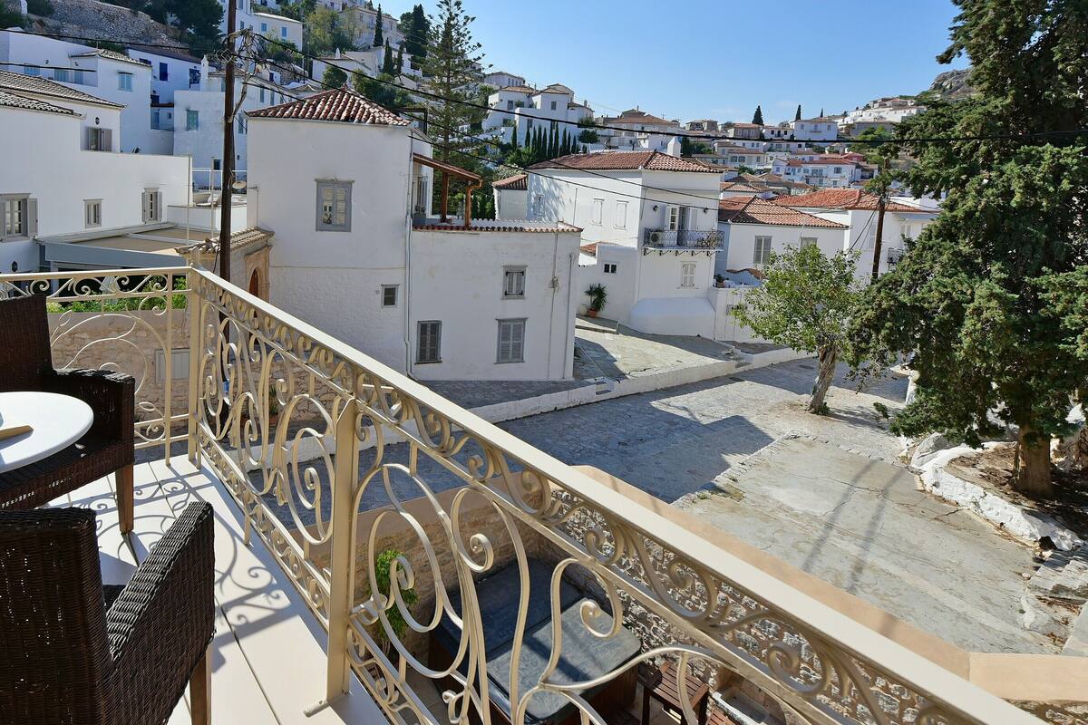 A balcony with wicker chairs and a table, overlooking a street with white buildings and a tree.